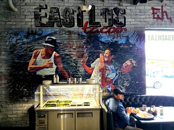 A mural over a salsa bar shows the illustrated figures of three young men from the film Blood In, Blood Out, with one holding another back and laughing while one stands in a fighting pose. A customer sits at a black booth his is tray of breakfast.