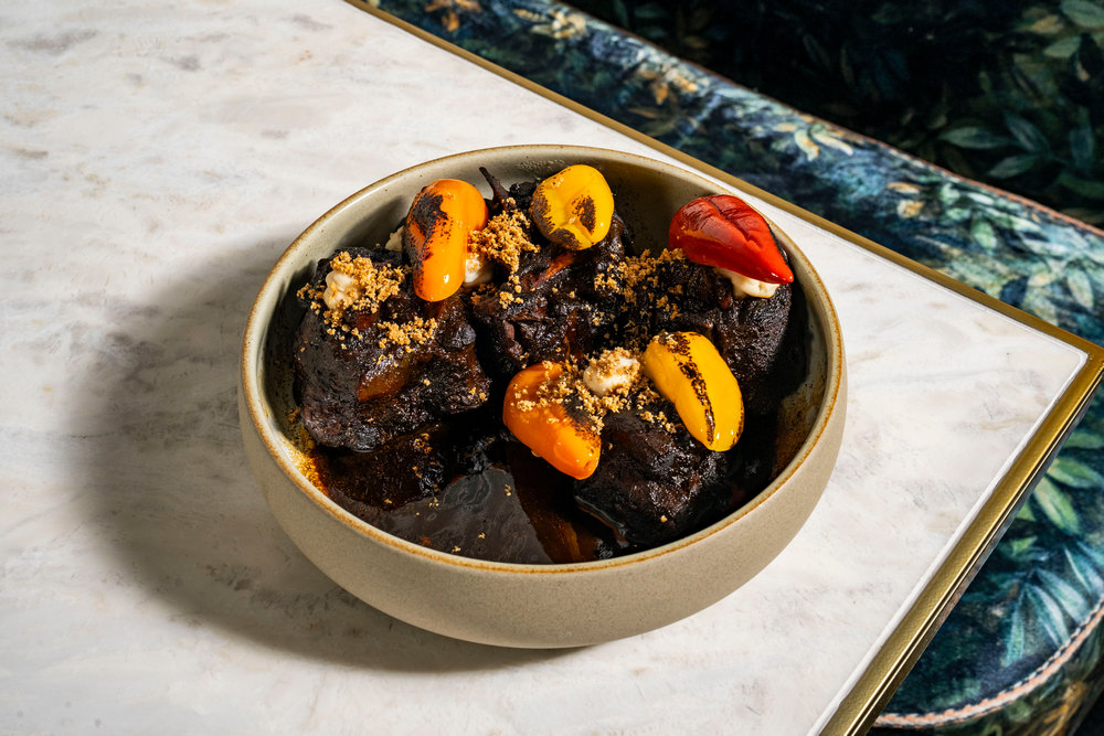 A tan bowl of black oxtail topped with red and yellow Fresno chiles on a marble tabletop.