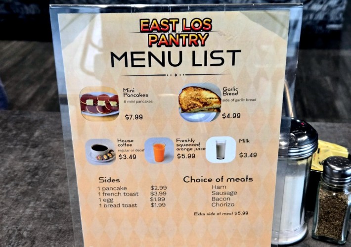 A tan menu in a plastic holder, on the tables at East Los Tacos, details some of the Original Pantry offerings there, including mini pancakes, garlic bread, house coffee, freshly squeezed orange juice, milk, ham, sausage, bacon, chorizo, French toast, eggs, and toast.