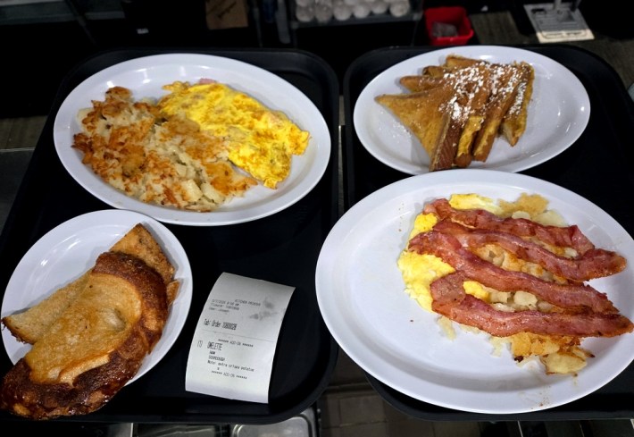 An order of different plates of potatoes, eggs, French toast, garlic bread, and bacon awaiting transfer to a table, on two trays.