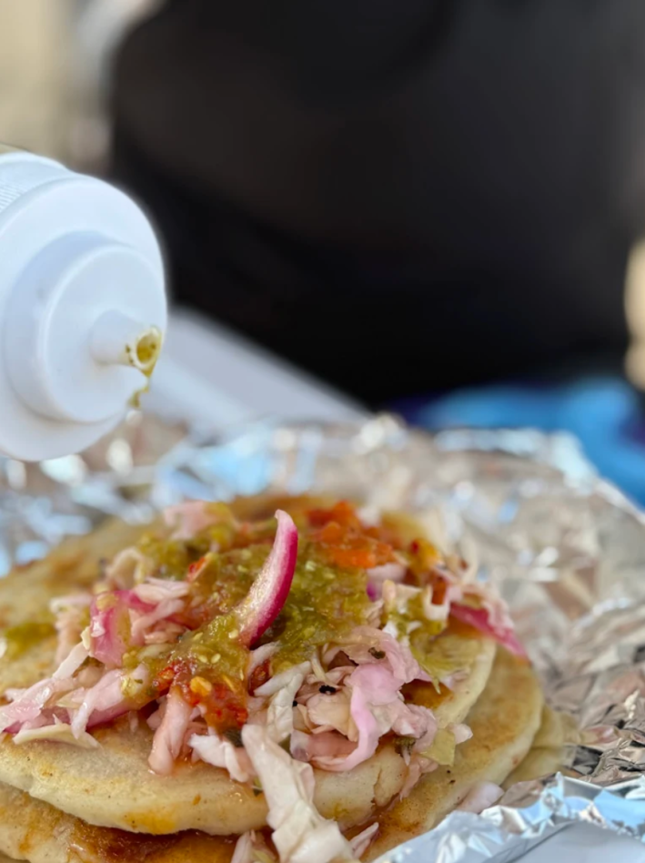 A pupusa preparada with curtido and topped with green salsa.