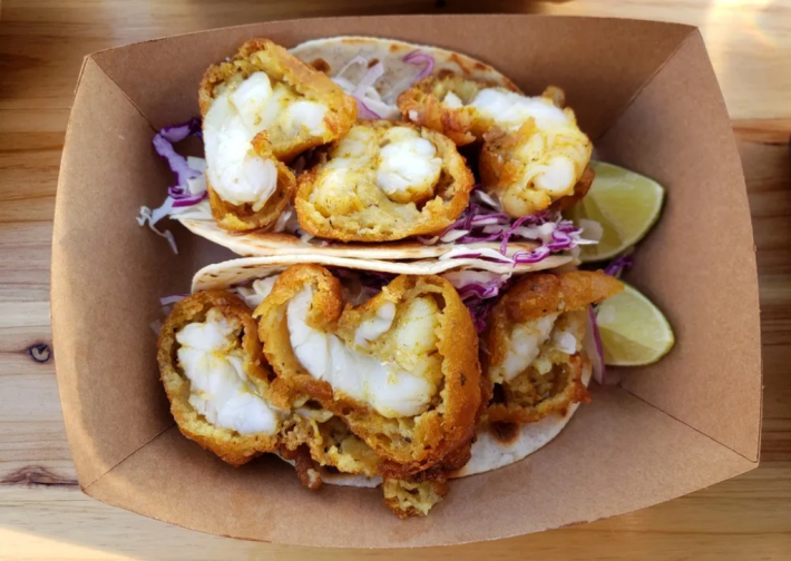 2 battered lobster tacos at Razos Fish Tacos in North Hollywood, in a brown cardboard box with limes on the side and the meat peaking out of the orange batter