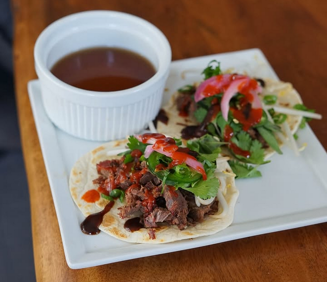 Two pho tacos on a white square plate with a ramekin of pho broth. The tacos are on flour tortillas and have beef, jalapeno, bean sprouts, cilantro, sriracha and hoison sauce.