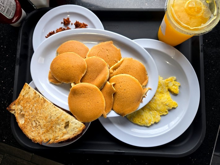 A tray with plates of mini-pancakes, garlic bread, chorizo, eggs, and orange juice at East Los Tacos.
