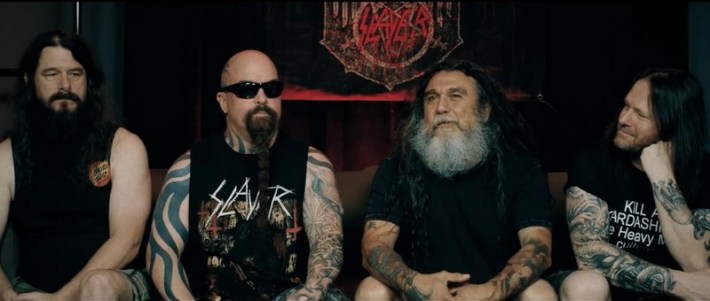 The four members of Slayer, seated and heavily tattooed in black t-shirts during an interview. Kerry King wears shades and a black tank top.