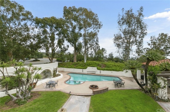 A uniquely shaped pool in back of a mansion, with pool furniture, a lawn, and green trees