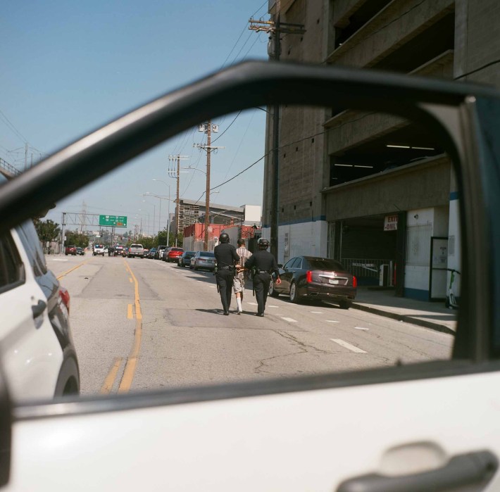 Two cops lead a person away in handcuffs, seen through the back, framed by a car window