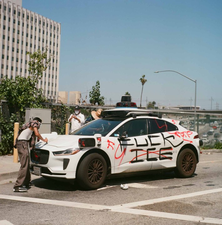 A white Waymo car spray-painted FUCK ICE, as a person writes on it and another takes a photo with their phone