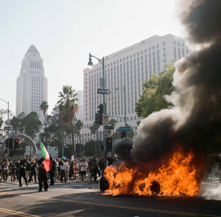 A car burns, releasing a lot of smoke, in front of Downtown LA buildings, with a crowd of protestors befor it