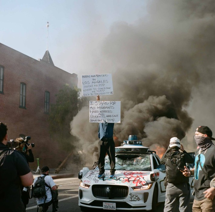 A person stands atop a trashed Waymo car holding protest signs, as smoke billows behind them