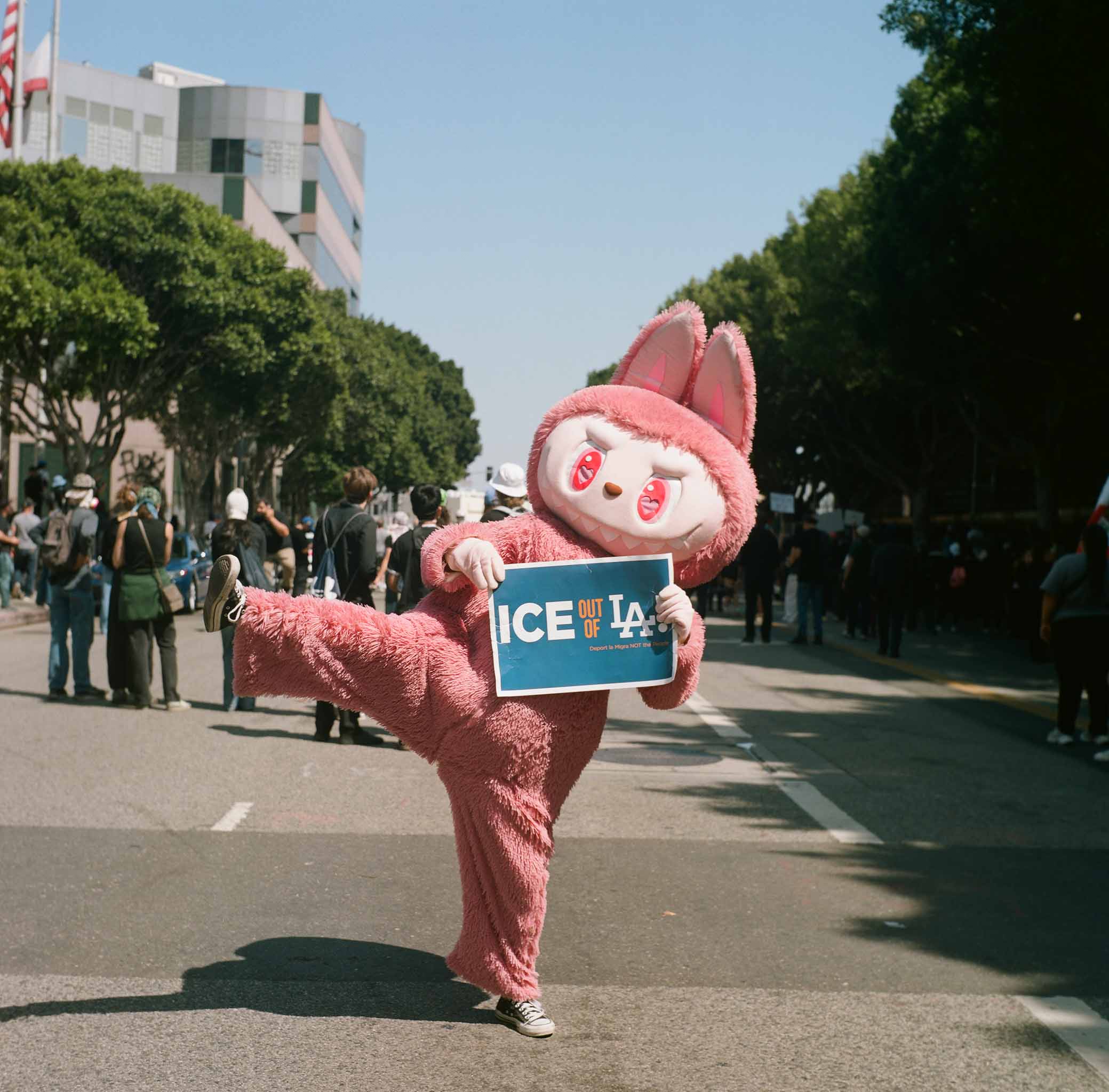 A person in a pink Labubu costume holding a sign that says "ICE Out of LA"