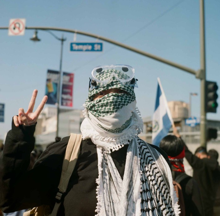 A protestor with a keffiyeh on their head holds up two fingers in a peace sign under the Temple St sign