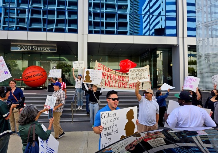 Protestors circling the walkway to a high-rise building holding up signs against ICE and mass surveillance