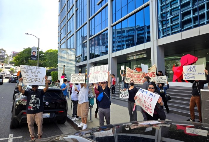 Protestors outside of a high-rise building holding up signs against ICE, the war on the poor, and mass surveillance