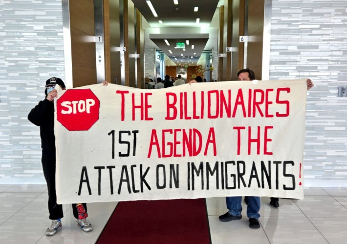A protest sign being held up by two people that says "The billionaires 1st agenda the attack on immigrants"
