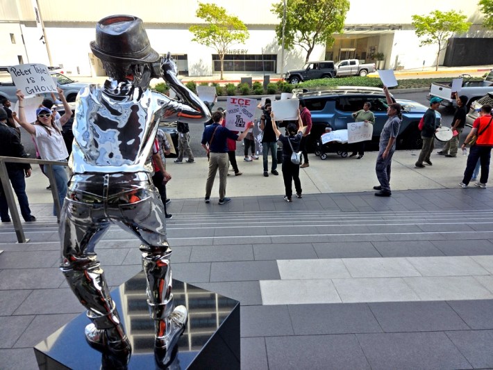 A metal statue of a photographer faces a crowd of people protesting on a sidewalk