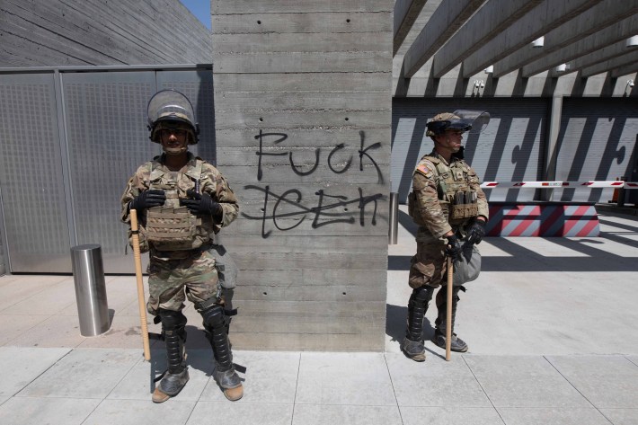 Two uniformed soldiers stand with a pillar in between that says FUCK ICE, with the ICE crossed out