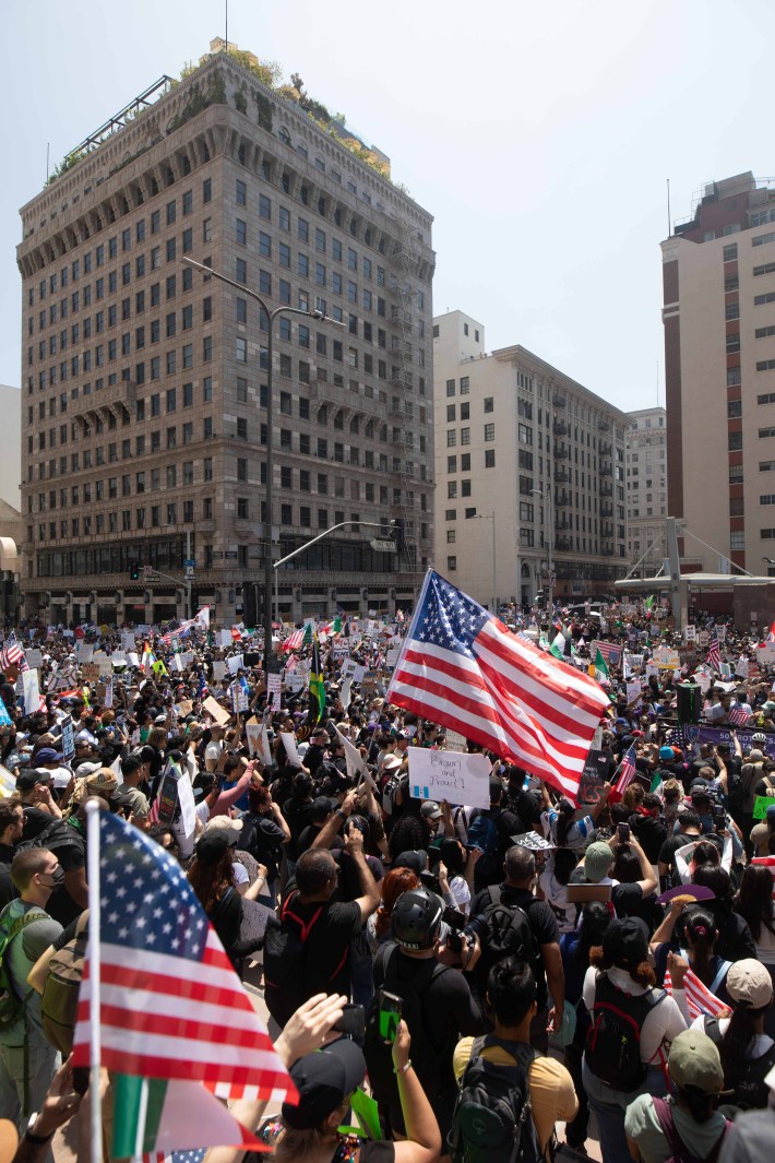 A crowd of protestors in Downtown LA, with one U.S. flag waving in front of tall buildings