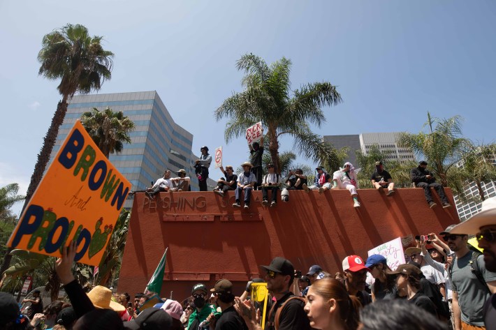 A crowd sits atop a sculture at LA's Pershing Square while an orange sign is upheld saying "Brown and Proud"