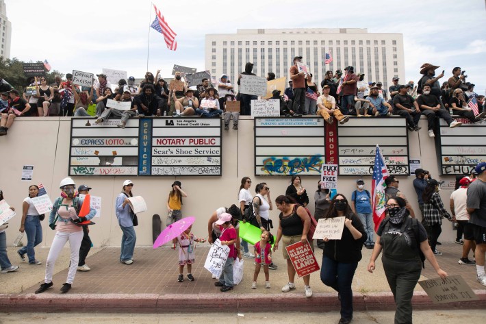 A crowd sits atop a wall in front of an US flag, while protestors and children stand by the curb below