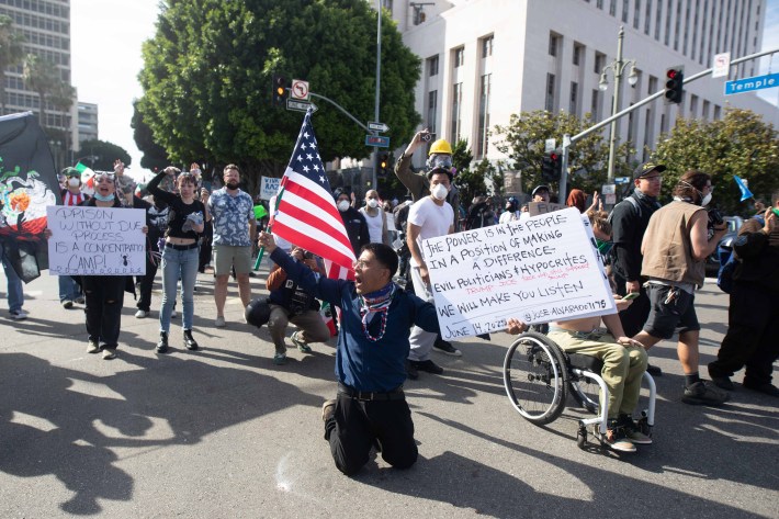 A protestor on his knees holding a US flag and a protest sign as others stand behind him
