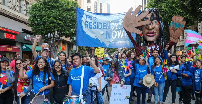 Activists with Carecen in blue shirts walking down the street, some beating drums, one holding a large puppet of a human head