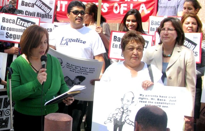 A woman speaks into amicrophone surrounded by demonstrators holding signs about former LA sheriff Baca