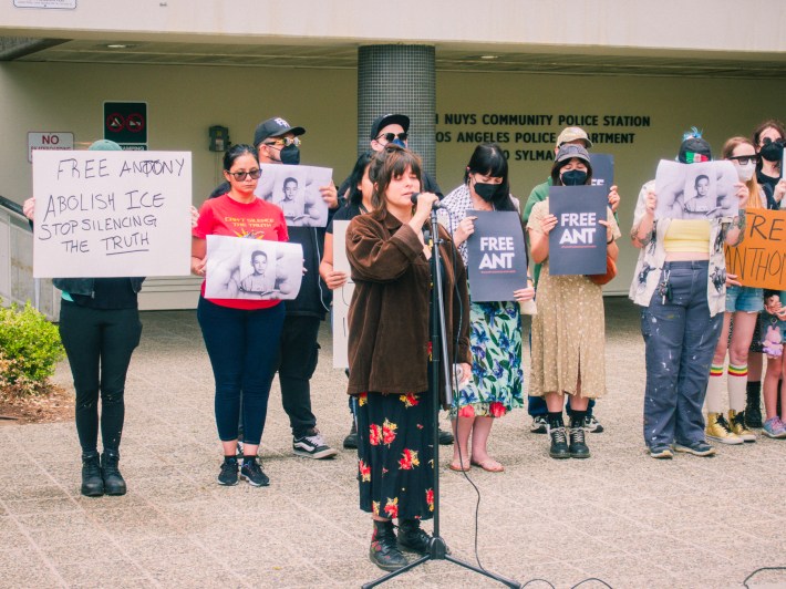A speaker with a microphone, speaking in support of jailed journalist Anthony Orendoff, with several more supporters behind them holding signs that say "Free Ant" and "Free Anthony Abolish ICE Stop Silencing the Truth"