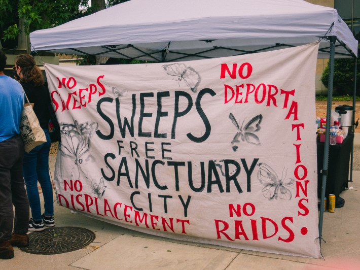 A large sign with black and red writing that says "No Sweeps" "Sweeps Free Sanctuary City" "No Deportations" No Raids" and "No Displacement"