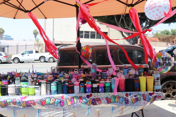 A street vendors stand decorated for her birthday her stand filled with reusable water bottles covered in balloons and pink party strands.
