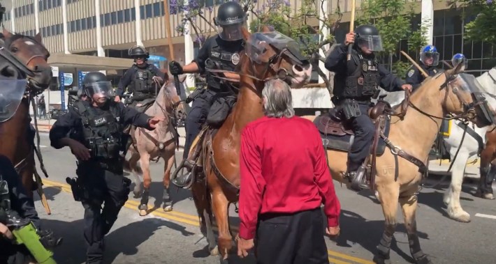A 77-year-old man in a red shirt is seen from behind as he is confronted by a mounted police officer, with other officers both on horses and on foot next to them