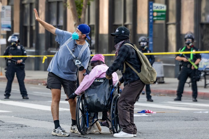 A woman in a wheelchair is assisted by two protestors while police in riot gear watch in the background.