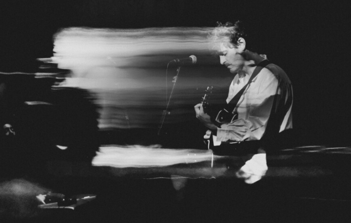 With blurry tracers extending from his body, a man plays a guitar in black and white