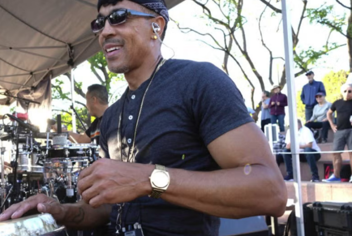 A man in sunglasses and a black t-shirt plays a hand drum