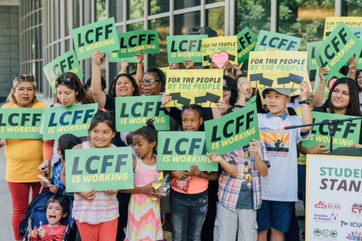 A group of children hold up green signs that say LCFF is working
