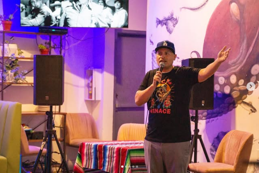 A comedian in a black t-shirt and Dodgers cap speaks into a microphone onstage while holding one hand out to his side