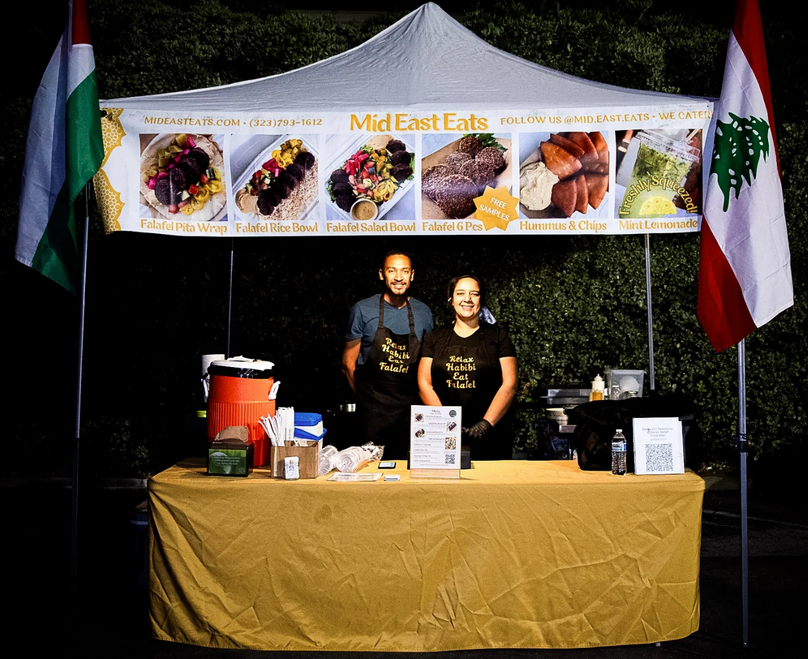 A smiling couple stands in front of their food stand flanked by the flags of Lebanon and Palestine.
