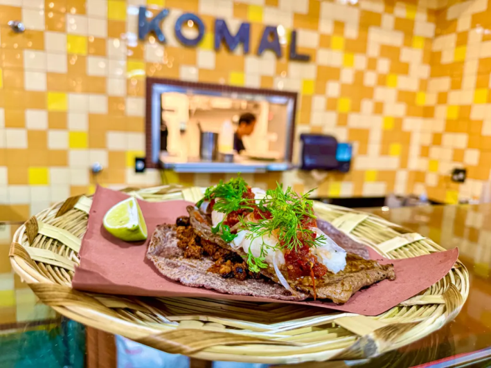 A straw plate with a taco on ablue corn tortilla in its middle, topped with microgreens and a lemon the side. A window in back has a sign that says KOMAL above it