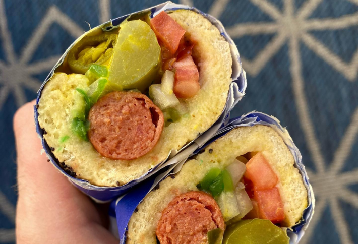 A Chicago-style hot dog, seen in the cross-section, with green relish, onions, a pickle spear, sliced tomatoes, sport pepper, yellow mustard, and celery salt.