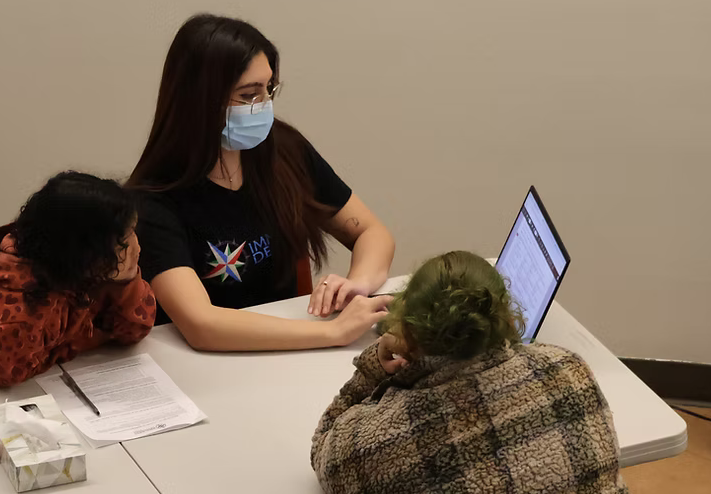 A woman in a mask shows a woman and a child something on a laptop
