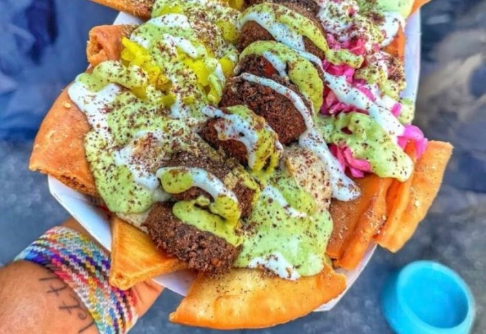 A bed of pita chips topped with falafel and sauces being held in a hand attached to a wrist with a lot of bracelets