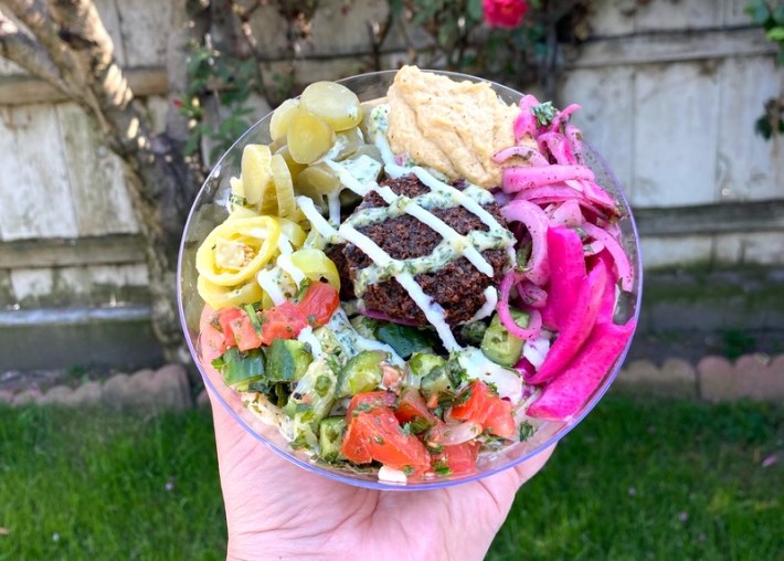 A colorful plate with falafel in the center crisscrossed with a white sauce and surrounded with salad, hummus, and pickled vegetables