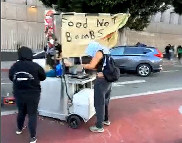 A screenshot of a video showing a flag that says Food Not Bombs over a couple of people preparing tacos on a cart