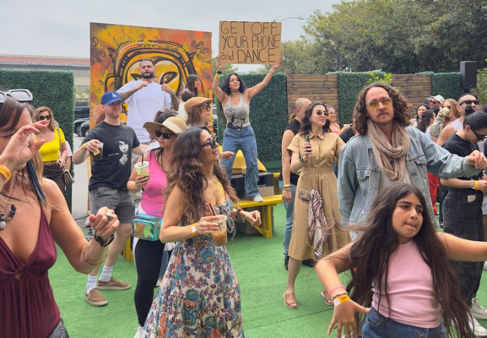 A group of people dance at a morning rave. One holds a sign that reads: "GET OFF YOUR PHONE AND DANCE"