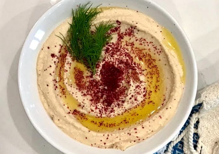 A white bowl full of hummus topped with olive oil, dill, and a red ground spice.