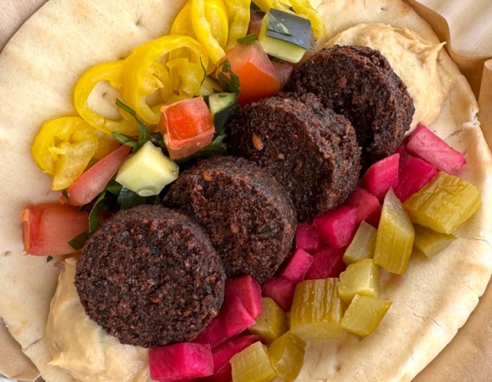 An open pita with four falalfel balls, over hummus and with pickled radish, tomato, celery, onion, and peppers on the side