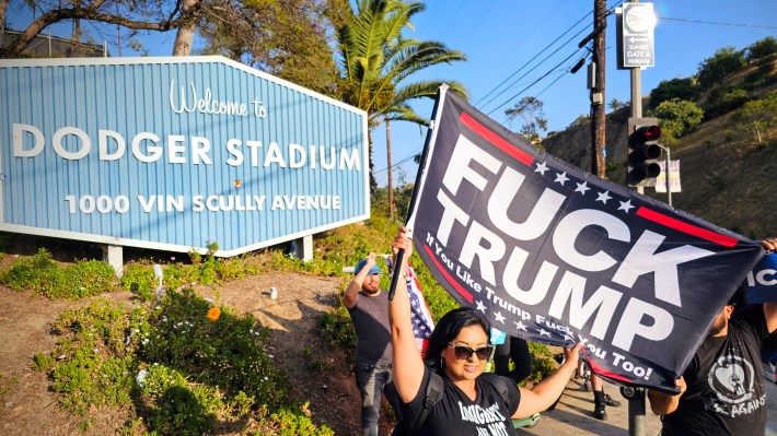 A protester holds a "FUCK TRUMP" flag outside of Dodger Stadium. The iconic "Welcome to Dodger Stadium" sign is seen in the background.
