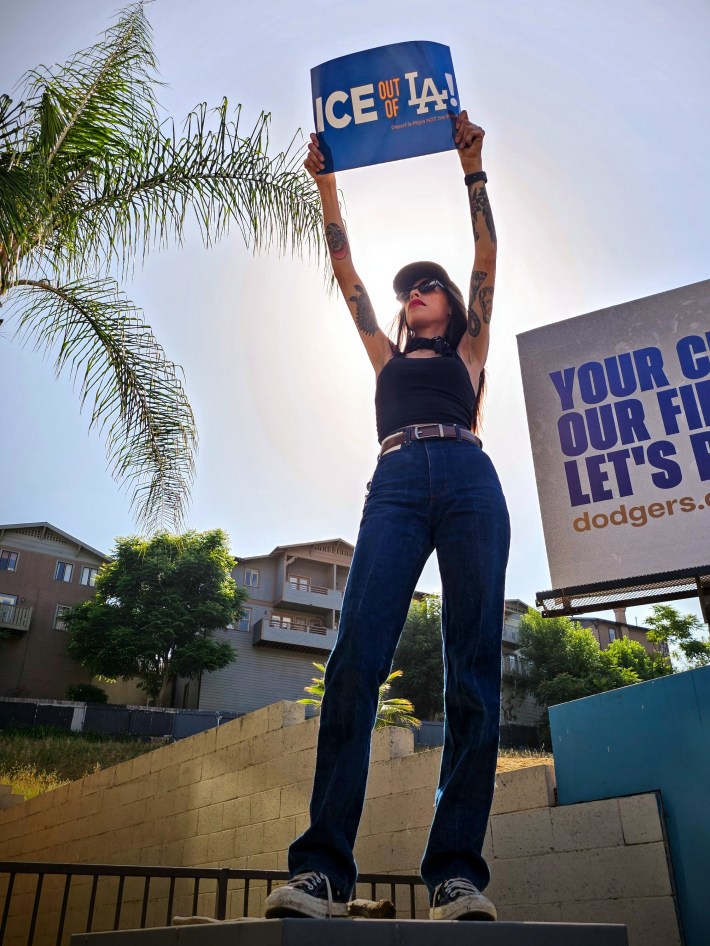 A woman protester holds an "ICE out of LA" sign while standing on a utility box.