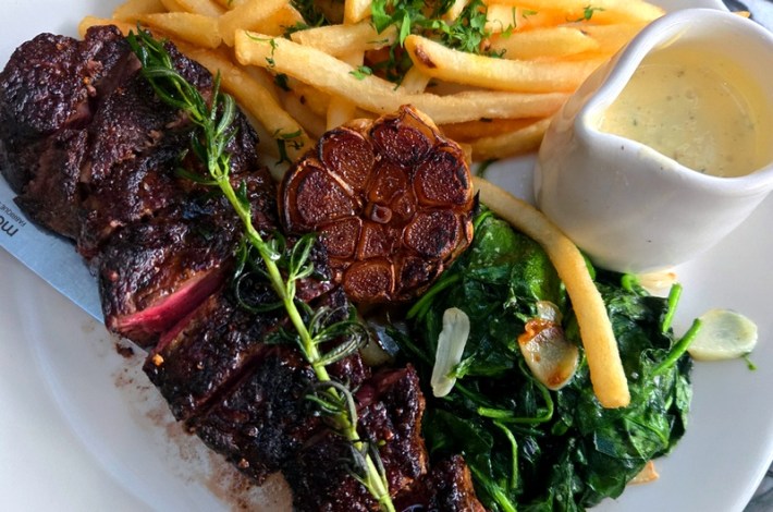 A sliced steak topped with rosemary and a cooked bulb of garlic, aside fries and a small white vase of yellow sauce
