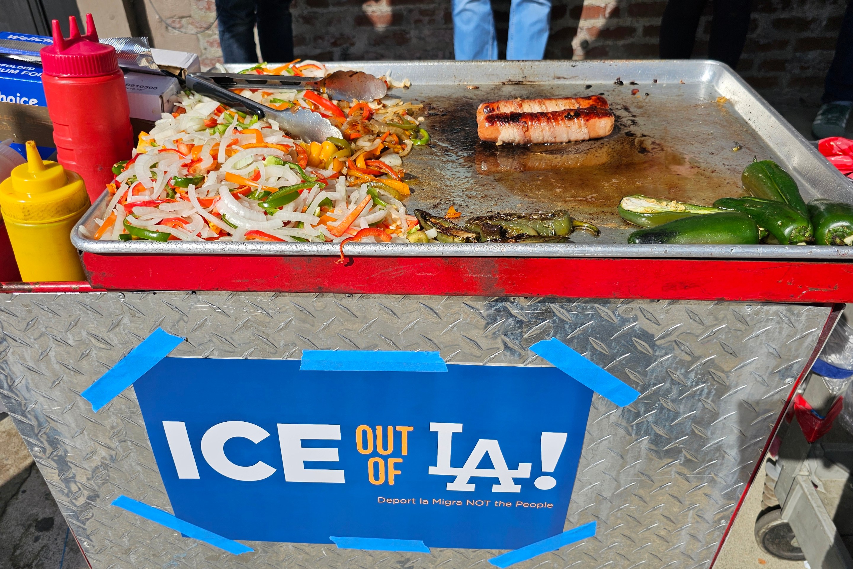 A street vending cart topped with bacon wrapped hotdogs and grilled veggies. There is an "ICE out of LA!" poster on the front of the cart.